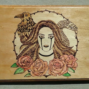 May include: Wooden plaque featuring a pyrography design. The artwork depicts a woman with long hair, a raven, and a snake within a circular frame. Pink roses with green leaves are at the bottom. The design is burned into the wood.