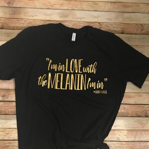 May include: Black t-shirt with gold lettering that reads "I'm in love with the MELANIN I'm in" with the words "Love" and "MELANIN" in a larger font. The text is centered on the shirt.