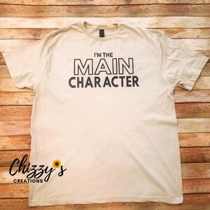May include: A white t-shirt with black text that reads "I'm the MAIN CHARACTER". The shirt is on a wooden background.