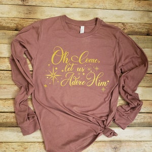 May include: Mauve long-sleeve shirt with the text "Oh Come, let us Adore Him" in gold script. The design includes a star and starbursts. The shirt is knotted at the waist and has a crew neck.