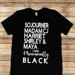 May include: Black t-shirt with white text that reads "Sojourner Madam CJ Harriet Shirley & Maya I am Phenomenally BLACK"