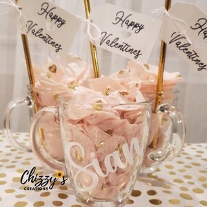 May include: Three glass mugs filled with pink and gold confetti, each with a gold straw and a white tag that says "Happy Valentines". The mug in the center has the name "Sian" written on it.