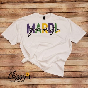 May include: White t-shirt with the words "MARDI GRAS" in purple, green, and yellow, with a black cursive font overlay. The shirt is knotted at the bottom. The brand "Chizzy's Creations" is printed in black cursive font.