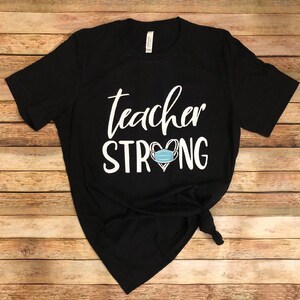 May include: Black t-shirt with white text that reads "teacher strong" with a heart containing a blue face mask.