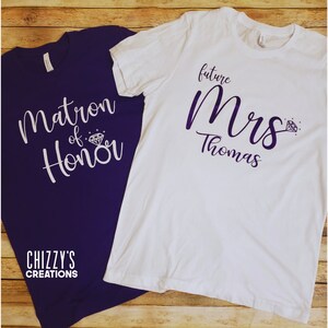 May include: Two t-shirts, one purple and one white, with text printed on them. The purple shirt says "Matron of Honor" with a diamond ring graphic. The white shirt says "future Mrs. Thomas" with a diamond ring graphic.