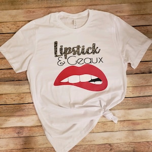 May include: White t-shirt with a red lip graphic and the text "Lipstick & Geaux" in black with a leopard print pattern.