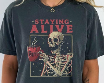 Może przedstawiać: Czarny T-shirt z grafiką przedstawiającą szkielet trzymający kubek z kawą. Tekst "STAYING ALIVE" jest nadrukowany nad szkieletem.