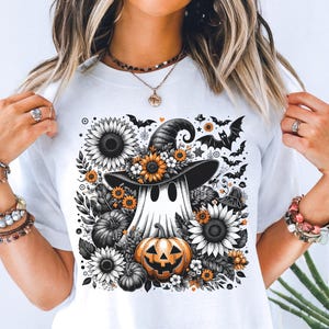 Puede incluir: Camiseta blanca con un diseño de Halloween. El diseño presenta un fantasma con un sombrero de bruja, rodeado de girasoles, calabazas y murciélagos. El fantasma tiene ojos negros y una calabaza. El diseño utiliza negro, blanco y naranja.