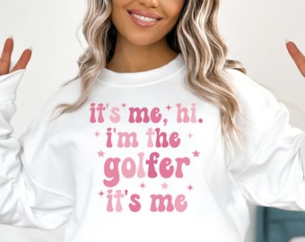 以下が含まれることがあります： ピンクの文字で「it's me, hi. i'm the golfer it's me」と書かれた白いスウェットシャツ。文字の周りにピンクの星が散りばめられています。