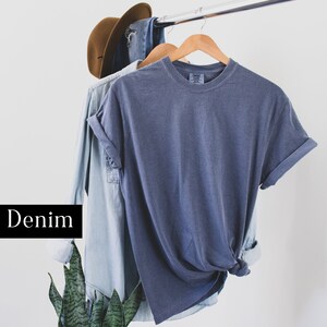 Pode incluir: Uma camiseta de brim azul com mangas dobradas pendurada em um cabide de madeira. A camiseta est&aacute; pendurada sobre uma camisa de brim azul claro.