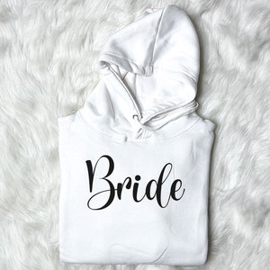 以下が含まれることがあります： 白いフード付きスウェットシャツに、黒い筆記体で「Bride」とプリントされています。