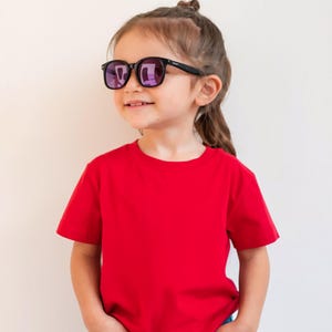 May include: A young child wearing black-framed sunglasses with purple lenses and a red t-shirt. The child is smiling and looking to the side. The background is a plain, off-white wall.