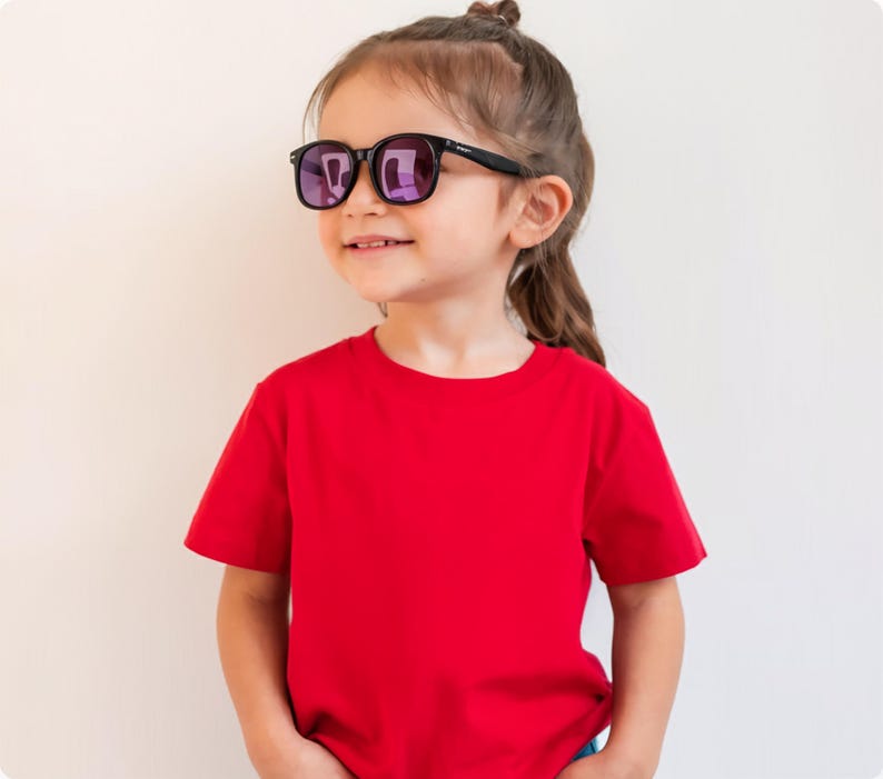 May include: A young child wearing black-framed sunglasses with purple lenses and a red t-shirt. The child is smiling and looking to the side. The background is a plain, off-white wall.