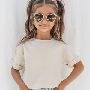 May include: A young person wearing heart-shaped sunglasses with a light pink frame. The person is wearing a cream-colored, short-sleeved t-shirt with rolled-up sleeves. The background is a plain white surface.
