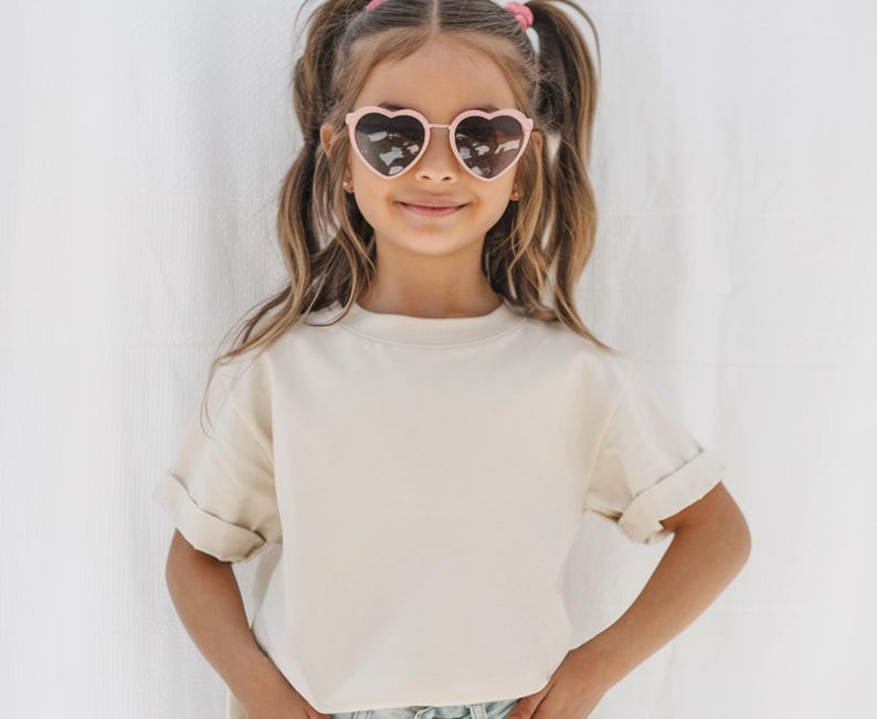 May include: A young person wearing heart-shaped sunglasses with a light pink frame. The person is wearing a cream-colored, short-sleeved t-shirt with rolled-up sleeves. The background is a plain white surface.