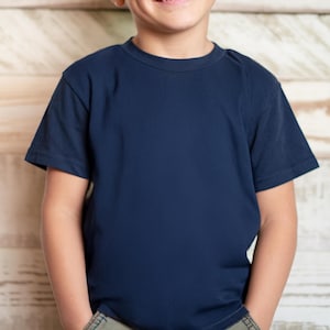 May include: A navy blue crew neck t-shirt. The short-sleeved shirt is made of a soft material. The shirt is shown on a person in front of a light brown wooden background.