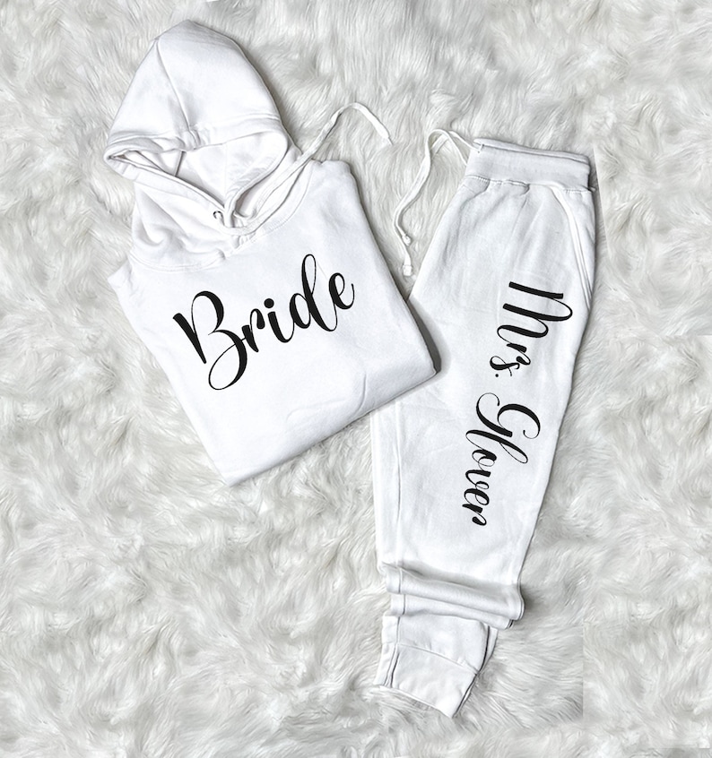 以下が含まれることがあります： 白いフード付きスウェットシャツに「Bride」の文字がプリントされ、白いスウェットパンツに「Mrs. Glover」の文字が側面にプリントされています。