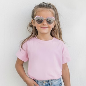 May include: A young person wearing heart-shaped sunglasses with a pink frame and a light pink t-shirt. The person is smiling and wearing blue jeans. The background is a white wall.