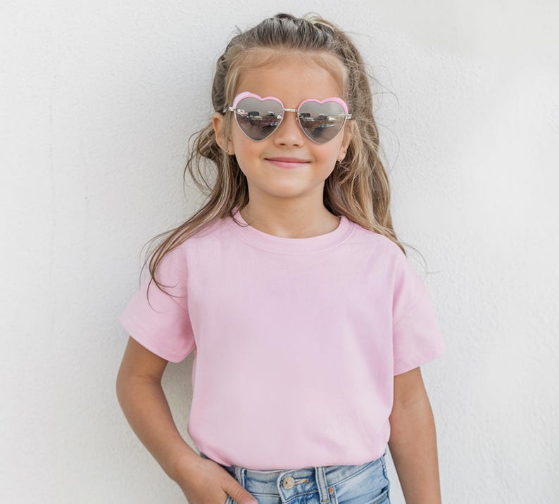 May include: A young person wearing heart-shaped sunglasses with a pink frame and a light pink t-shirt. The person is smiling and wearing blue jeans. The background is a white wall.