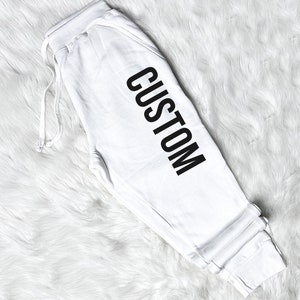 以下が含まれることがあります： 黒で「CUSTOM」とプリントされた白いスウェットパンツです。
