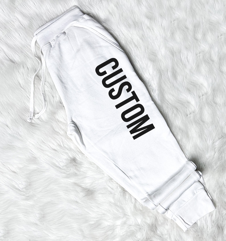 以下が含まれることがあります： 黒で「CUSTOM」とプリントされた白いスウェットパンツです。