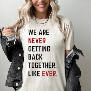 May include: Cream-colored t-shirt with the text "WE ARE NEVER GETTING BACK TOGETHER. LIKE EVER." in black and red lettering. The shirt is being worn by a person with blonde hair and a denim jacket.
