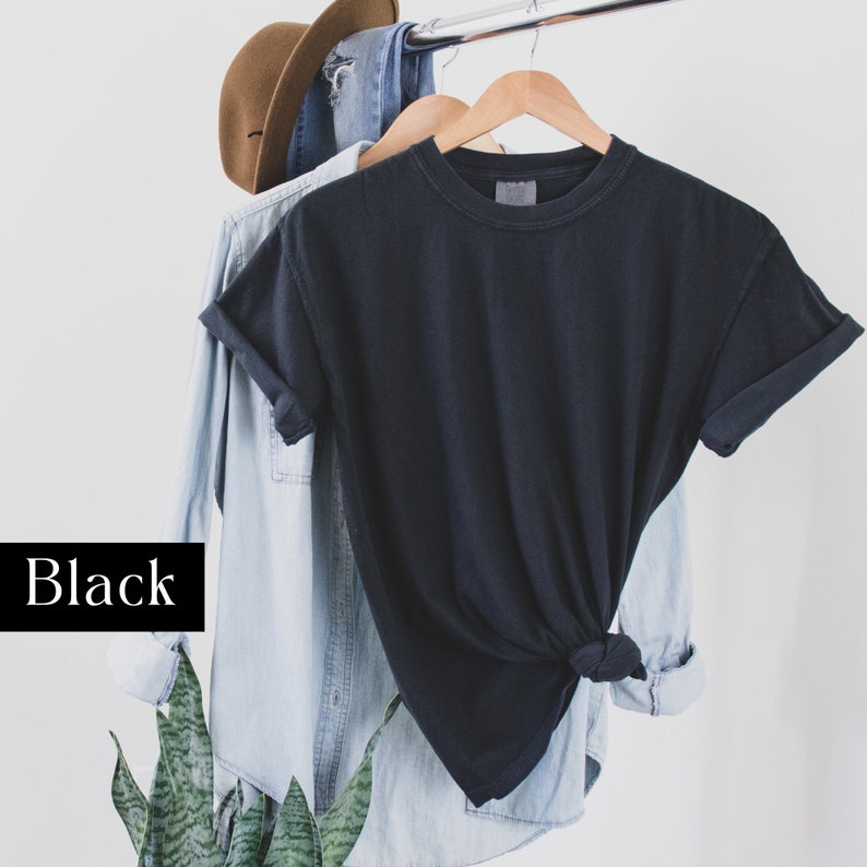 Pode incluir: Uma camiseta preta pendurada em um cabide de madeira. A camiseta est&aacute; dobrada e amarrada na parte inferior. A camiseta est&aacute; pendurada na frente de uma camisa de brim azul claro.