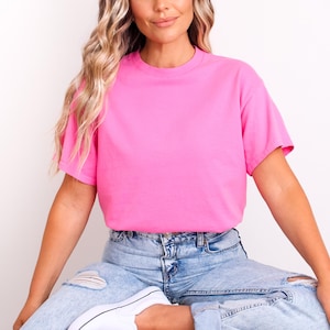 Op de afbeelding: Een felroze T-shirt met ronde hals. De top met korte mouwen wordt gecombineerd met lichtblauwe jeans met distressed details. Witte sneakers maken de casual outfit compleet. Het model heeft lang blond haar.