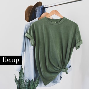 Pode incluir: Uma camiseta verde de c&acirc;nhamo pendurada em um cabide com uma camisa jeans azul por baixo. A palavra "C&acirc;nhamo" est&aacute; escrita em preto sobre um fundo branco.