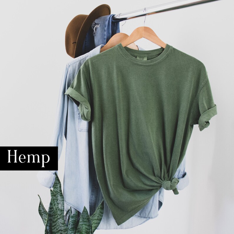 Pode incluir: Uma camiseta verde de c&acirc;nhamo pendurada em um cabide com uma camisa jeans azul por baixo. A palavra "C&acirc;nhamo" est&aacute; escrita em preto sobre um fundo branco.