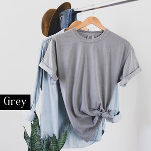 Pode incluir: Uma camiseta cinza pendurada em um cabide de madeira. A camiseta est&aacute; amarrada na parte inferior e tem as mangas dobradas.