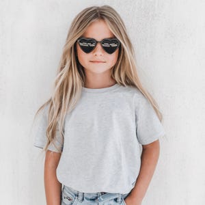May include: A young person wearing heart-shaped sunglasses with black frames. The person is wearing a light gray t-shirt and blue jeans. The background is a plain white wall.