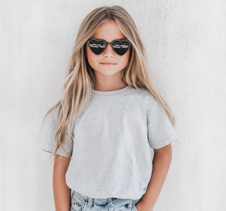 May include: A young person wearing heart-shaped sunglasses with black frames. The person is wearing a light gray t-shirt and blue jeans. The background is a plain white wall.