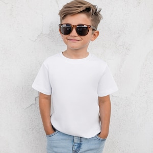May include: A young person wearing brown-framed sunglasses, a white t-shirt, and blue jeans. The person is standing in front of a textured white wall. The sunglasses have dark lenses.