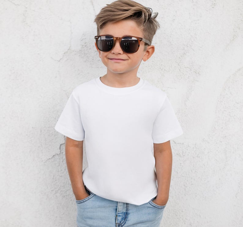 May include: A young person wearing brown-framed sunglasses, a white t-shirt, and blue jeans. The person is standing in front of a textured white wall. The sunglasses have dark lenses.