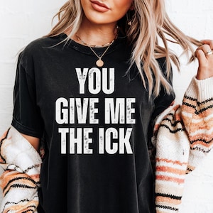 Puede incluir: Camiseta negra con el texto blanco "YOU GIVE ME THE ICK". La camiseta tiene un aspecto vintage y desgastado. La modelo lleva un collar dorado y un suéter a rayas. La camiseta es de color oscuro.