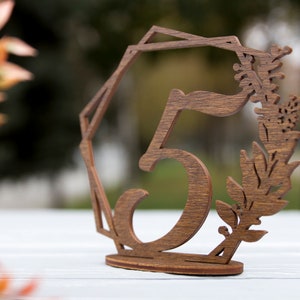 May include: Wooden table number with the number 5 cut out in the center. The number is surrounded by a geometric design and a floral design.