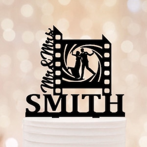 May include: Black silhouette of a couple dancing inside a film reel frame with the text "Mr. & Mrs. SMITH" on a white wedding cake decorated with white roses and green leaves.
