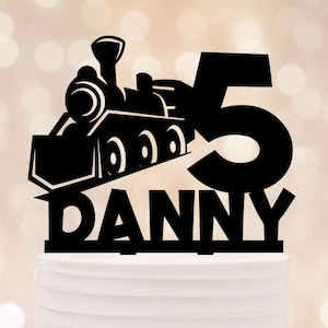 Könnte beinhalten: Schwarzer Tortenaufsatz mit einem Zug, der Zahl 5 und dem Namen "DANNY". Der Zug hat detaillierte Räder und einen Schornstein. Dieser Aufsatz ist für eine Geburtstagstorte gedacht.