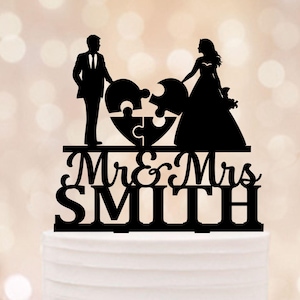Op de afbeelding: Een zwarte taarttopper met silhouetten van bruid en bruidegom, die een hartvormige puzzel vormen. De tekst "Mr & Mrs SMITH" staat eronder. De topper staat op een witte taart.