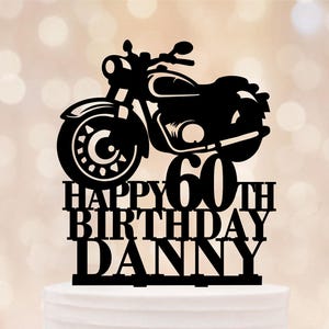 May include: Black motorcycle cake topper with the text "HAPPY 60TH BIRTHDAY DANNY". The topper is placed on top of a white frosted cake, with a blurred background.