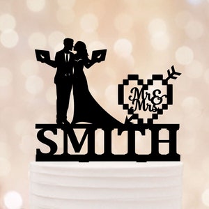 Peut inclure: Décoration de gâteau de mariage noire représentant une silhouette d'un couple s'embrassant tenant des tablettes, avec un cœur pixelisé et un motif de flèche indiquant "Mr & Mrs". Le nom de famille "Smith" est affiché en dessous.