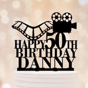 May include: Black cake topper with a film reel and movie camera design. The text reads "HAPPY 50TH BIRTHDAY DANNY". The topper is placed on a white cake with a blurred background.