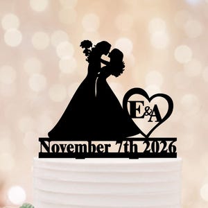 May include: Black silhouette cake topper featuring two brides embracing. A heart with the initials "E&A" is included. The date "November 7th 2026" is displayed below the figures. The topper is placed on a white cake.