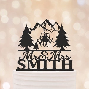 May include: Black cake topper featuring a mountain scene with skiers on a ski lift, pine trees, and the text "Mr & Mrs Smith". The topper is placed on a white frosted cake, perfect for a wedding or anniversary celebration.