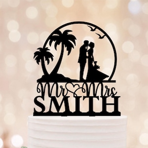 May include: Black wedding cake topper featuring a silhouette of a couple kissing under a circular frame with palm trees and birds. The words "Mr & Mrs SMITH" are displayed below the couple, perfect for a tropical themed wedding.