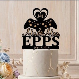 May include: A three-tiered wedding cake with gold accents and a decorative topper. The black topper features a heart, a game controller, the names "Mr & Mrs EPPS", and the date "July 11, 2026". A floral arrangement and a glass of champagne are also visible.