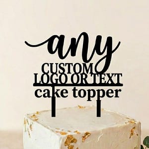 May include: A black cake topper featuring the text "any CUSTOM LOGO OR TEXT cake topper" in a mix of script and block fonts. The topper is inserted into a square white cake decorated with gold leaf flakes and a cluster of white roses and greenery.