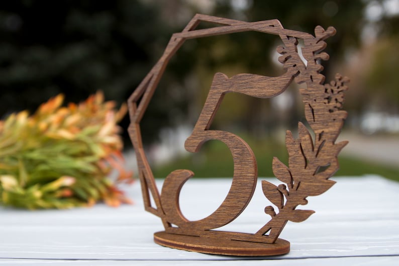May include: Wooden table number with the number 5 cut out and a floral design. The number is brown and the floral design is a light brown.