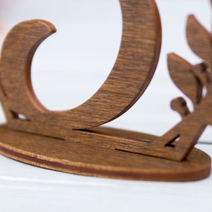 May include: A wooden table number holder with a decorative leaf design. The number is cut out of the wood and the holder has a rounded base.